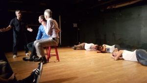 une scene de théâtre avec 2 personnes assisses sur une chaise, un homme debout et 3 personnes couchés